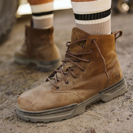 Person wearing 5.11 Tactical Rambler 6-inch Boots in Dark Coyote with striped crew socks, standing on a dirt track beside a muddy off-road vehicle tyre